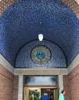 Two people are shown near the entrance of the Bird House at the Detroit Zoo in Royal Oak, Michigan. Pewabic tile in bright blues and golden iridescent line the domed entrance of the building. There is a prominent peacock motif lunette above the doorway completely made up of Pewabic tiles.