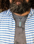 A man wearing a blue and white striped shirt paired with the Women's City Club necklace. The necklace features a rectangular metal pendant with a geometric design that includes teardrop shapes and smoothly curved lines in various shades of blue.