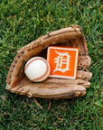 Baseball glove with a baseball and a Detroit Tigers tile in the grass.