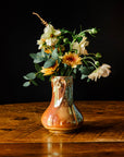 Sunstone Snowdrop vase with muted-yellow flowers and vibrant greenery on a wooden table against a dark background.