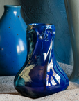 A handcrafted Azurite iridescent Pewabic Snowdrop vase is displayed in a beach setting that includes other Pewabic vases in Frost and Leland Blue in the background.