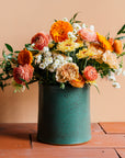 A colorful orange and blush flower arrangement in a Pewabic Crock featured in the matte Greenstone glaze, complete with visible iron flecks on its surface. The vessel is set upon terracotta roof tiles and a light peach background.