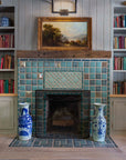 A fireplace with blue, green, and beige tiles, featuring architectural glazes, with bookshelves in the background and decorative blue and white vases on either side of the fireplace.