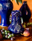 A handcrafted Azurite iridescent Pewabic Snowdrop vase is displayed on a table alongside fruits and in a setting that includes other Azurite vases in the background.