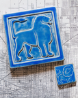 The Lion Tile featuring the Sky blue glaze sits next to a eight inch by eight inch right-facing Aztec Lion tile in the same glaze on a black and white map of Detroit.