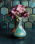 A Matte Green Iridescent Snowdrop vase with hellebore flowers on a dark slate foreground and green, tilled background.