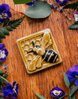 A glossy hand-painted Honeybee Tile in golden honey and black details surrounded by pansies, violets, and ferns.