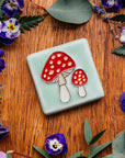 A hand-painted tile depicting a line drawing of a pair of Amanita mushrooms with classic red tops and white spots set on a light sea foam colored background glaze.