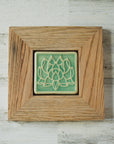 The Lotus tile features a line drawing of a blooming lotus flower, its pointed petals outstretching. It has a simple border around the design. A pale reclaimed wood frame holds the tile in the glossy pale blue Celadon glaze.