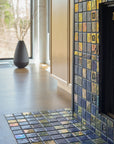 Angled photograph of a blue iridescent tiled fireplace. There is a matte charcoal vase in view. The fireplace is surrounded by light wood cabinets.