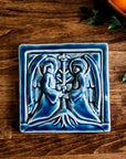 The square tile shows two angels in flowing robes with large feathered wings face each other, each has their hands grasping a bare tree growing between them. This tile features the glossy deep blue Ocean glaze.