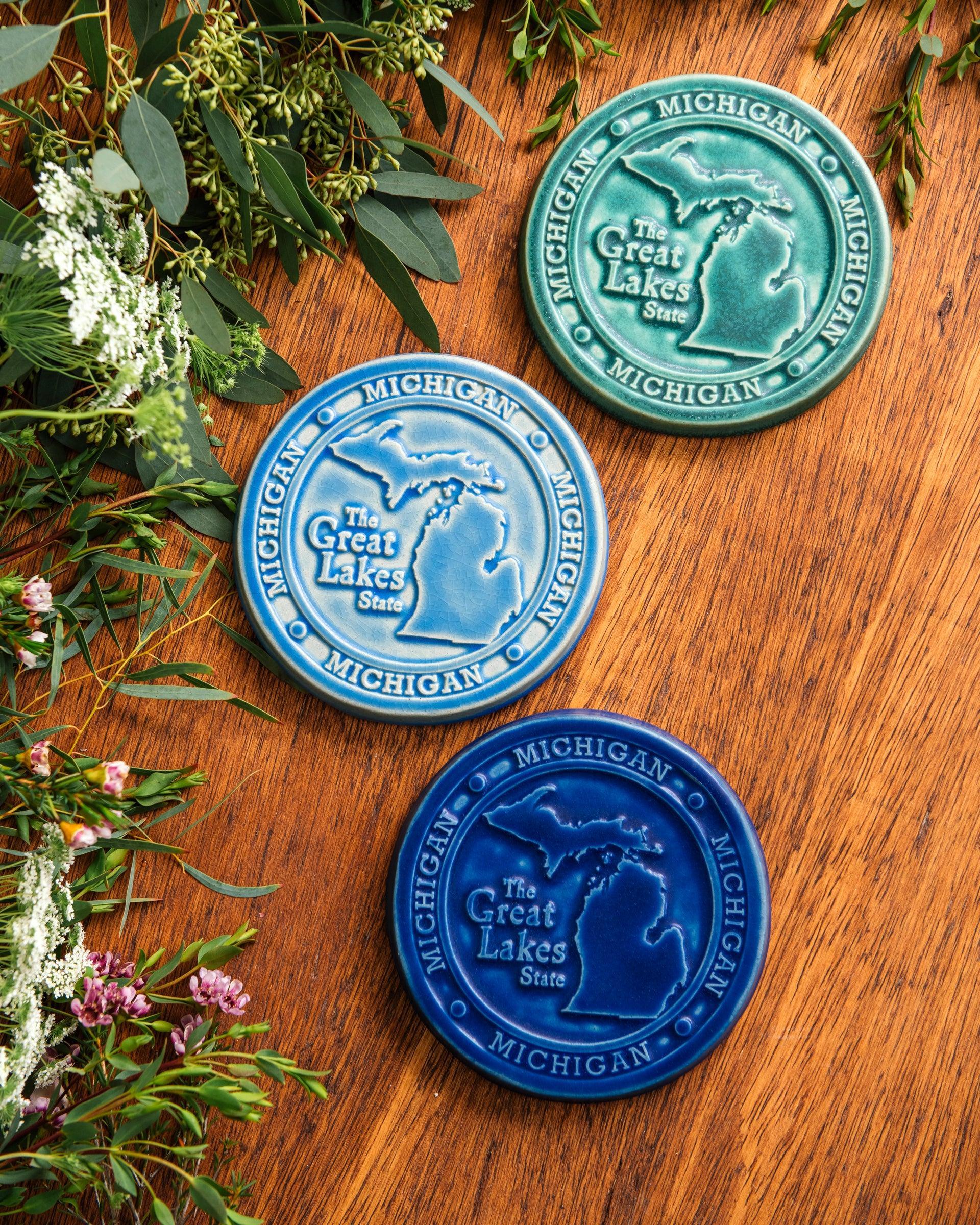 This round Michigan Trivet Tile features the outline of the state's two peninsulas including some of the larger northern islands of Michigan. To the left of the design reads "The Great Lakes State" and surrounding it, in a border, reads the word Michigan four times.