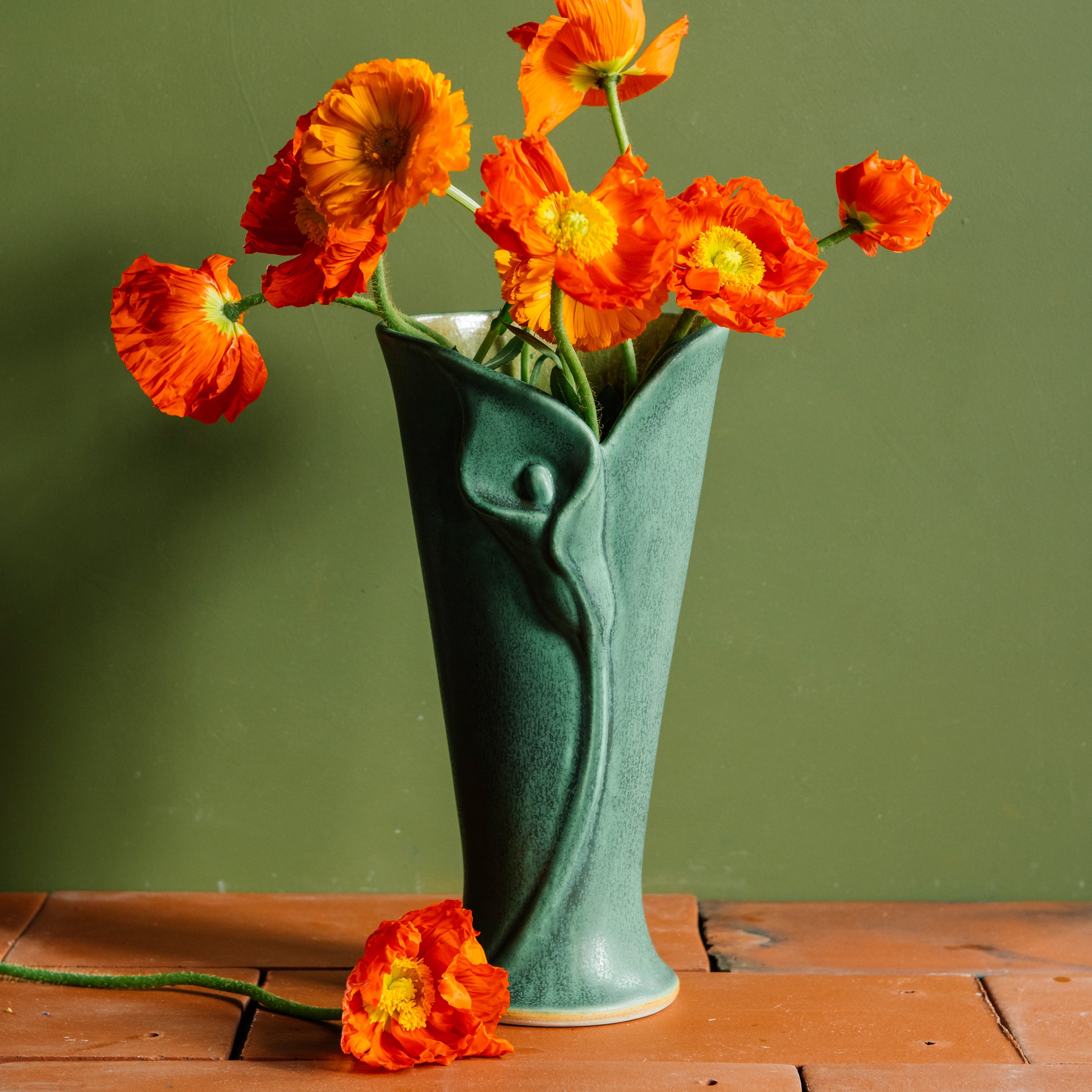Bright orange poppies whimsically positioned in varying directions inside a Pewabic Calla Vase. The vase is featured in a matte, textured, green "Elm" glaze and rests upon terracotta roof tiles with a verdant green background.  