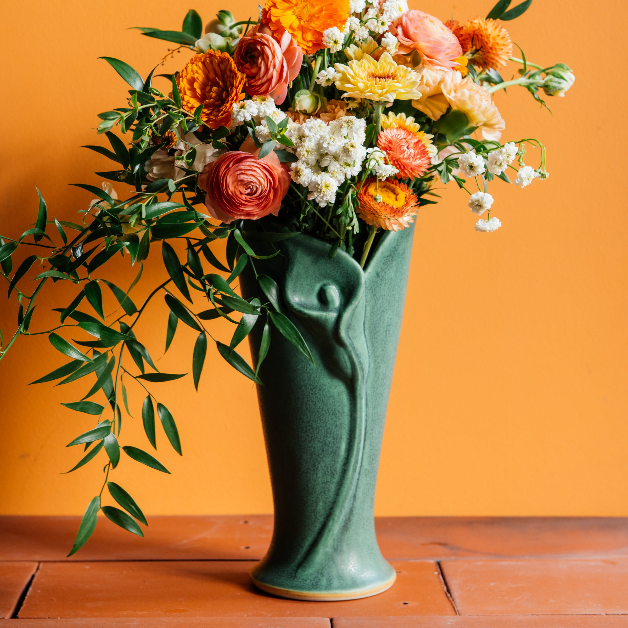 A dramatic bright orange, blush pink, and white floral arrangement with contrasting dark greenery spilling out of the top of a Pewabic Calla Vase in a matte Elm glaze. The vessel is set upon terracotta roof tiles against a bright orange backgrond. 