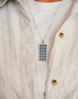 The Women's City Club necklace is a rectangular metal pendant with a geometric design that includes teardrop shapes and smoothly curved lines in various shades of blue. The necklace is worn by a person in a light-colored shirt with a linen button-up top.