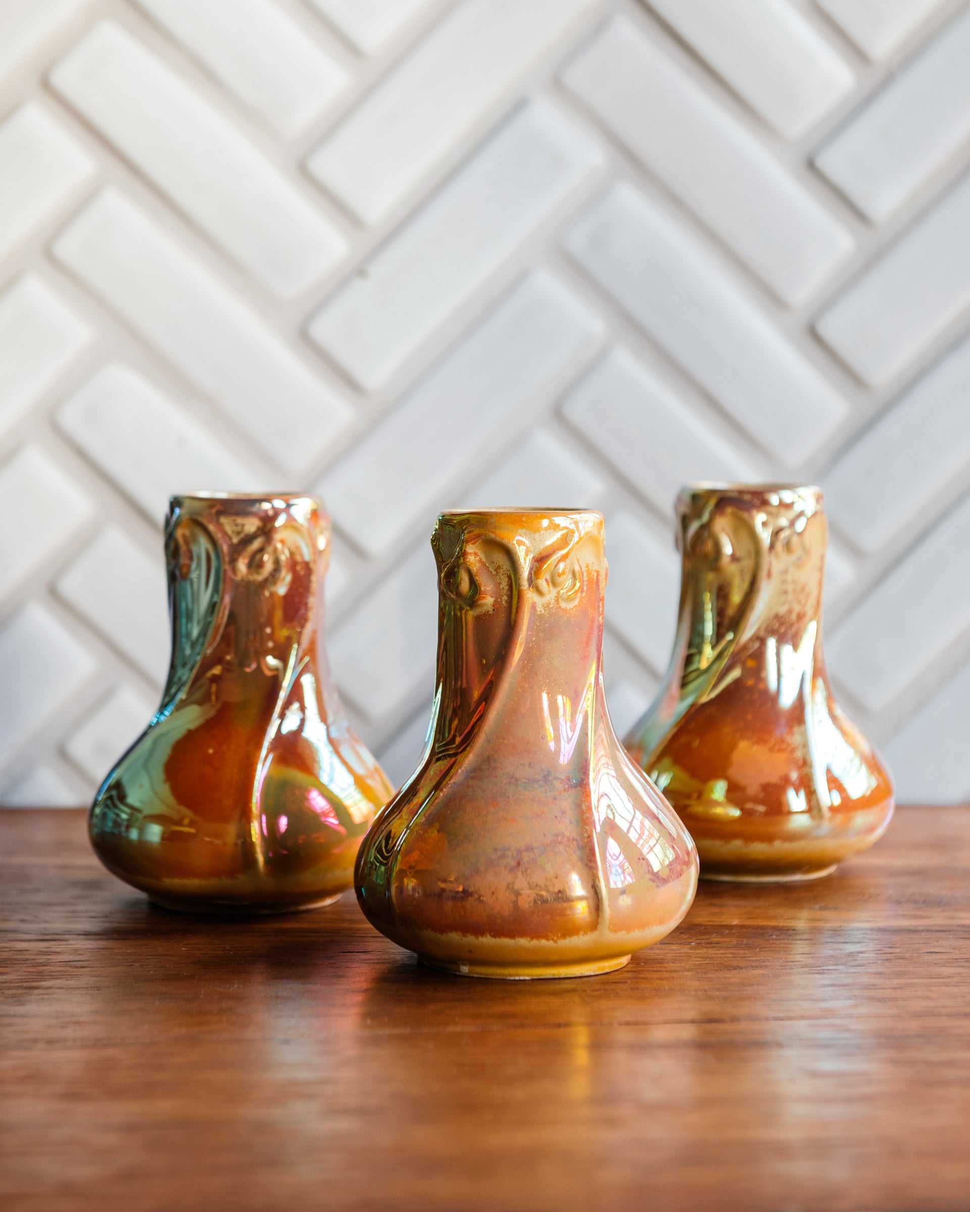 Sunstone Snowdrop vases, gleaming in bright iridescent yellow, pink, and orange tones set on a warm wooden foreground with a herringbone Pewabic tile pattern in a matte, white glaze. 