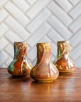 Sunstone Snowdrop vases, gleaming in bright iridescent yellow, pink, and orange tones set on a warm wooden foreground with a herringbone Pewabic tile pattern in a matte, white glaze. 