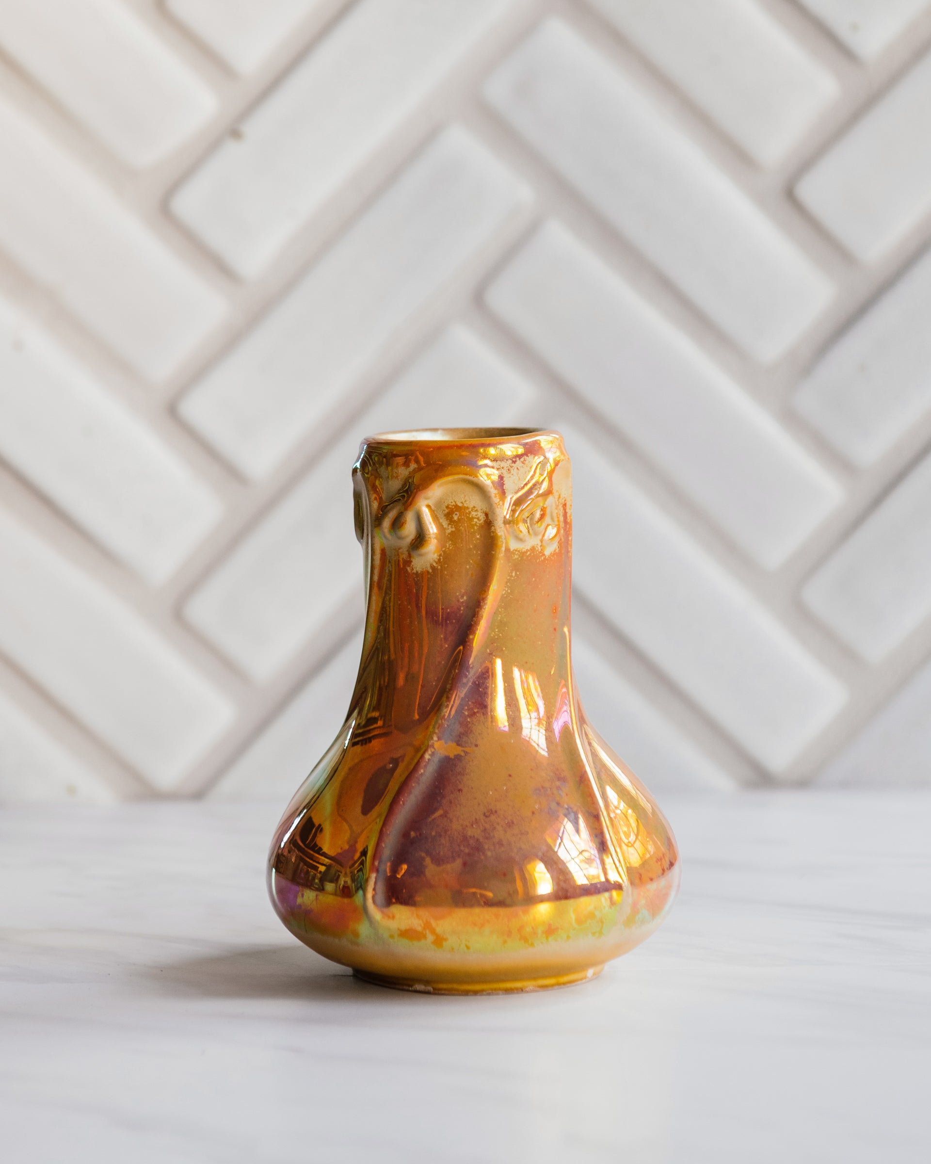 Sunstone Snowdrop vase, gleaming in bright iridescent yellow, pink, and orange tones set on white marble with a herringbone Pewabic tile pattern in a matte, white glaze. 