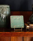 Decorative Old Detroit tiles in glossy Celadon glaze on a handcrafted Stickley cabinet next to a stack of books and a historically-inspired Pinecone Vase. 