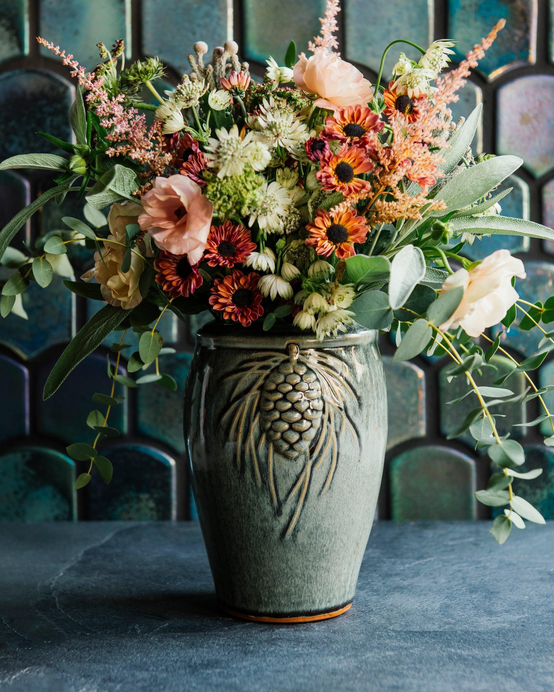 Glossy, deep green Balsam Pinecone vase with blush-pink and vibrant green flowers against an Aurora Iridescent tile wall. 