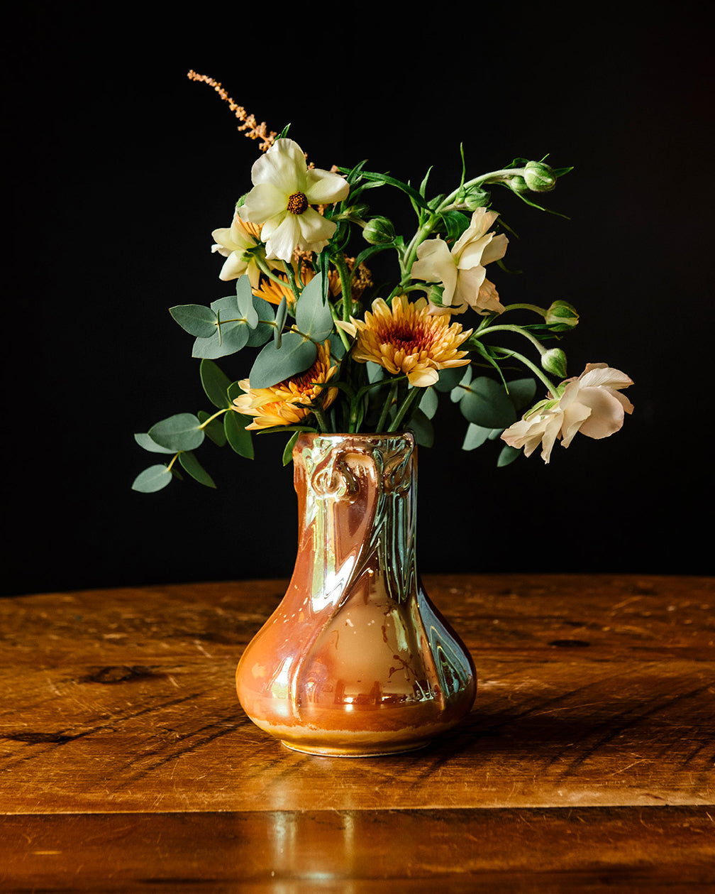 Sunstone Snowdrop vase with muted-yellow flowers and vibrant greenery on a wooden table against a dark background.