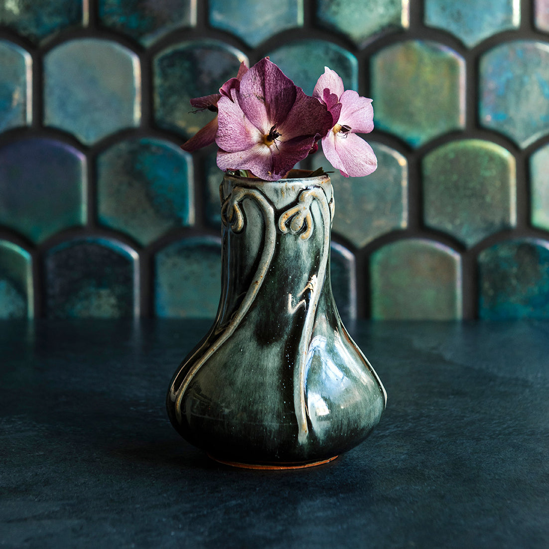 Glossy, deep green Balsam Snowdrop vase with purple hellebore flowers against an Aurora Iridescent tile wall. 