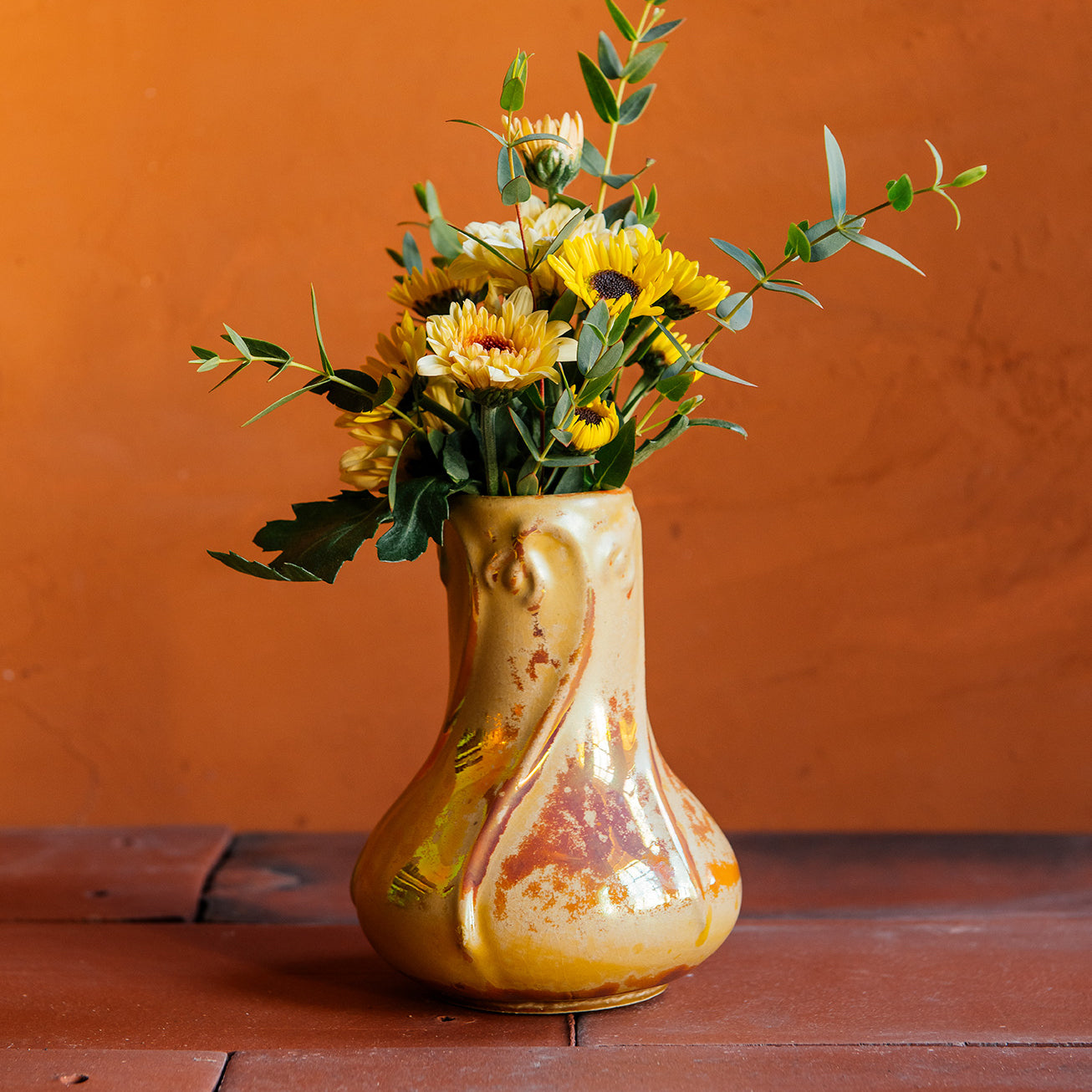 Delicate slip-casted snowdrop floral motifs dance across this Snowdrop Vase in a Sunstone Iridescent glaze that shimmers with the colors of a sunset. The texture of the vase varies with areas that are matte-yellow and reflective features glistening in orange and pinkish hues. The vase rests on terracotta roof tiles with a lime-wash orange background. Dainty yellow florals and dark, moody greenery make up a brilliant feature bouquet. 