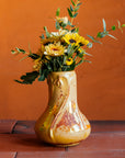 Delicate slip-casted snowdrop floral motifs dance across this Snowdrop Vase in a Sunstone Iridescent glaze that shimmers with the colors of a sunset. The texture of the vase varies with areas that are matte-yellow and reflective features glistening in orange and pinkish hues. The vase rests on terracotta roof tiles with a lime-wash orange background. Dainty yellow florals and dark, moody greenery make up a brilliant feature bouquet. 