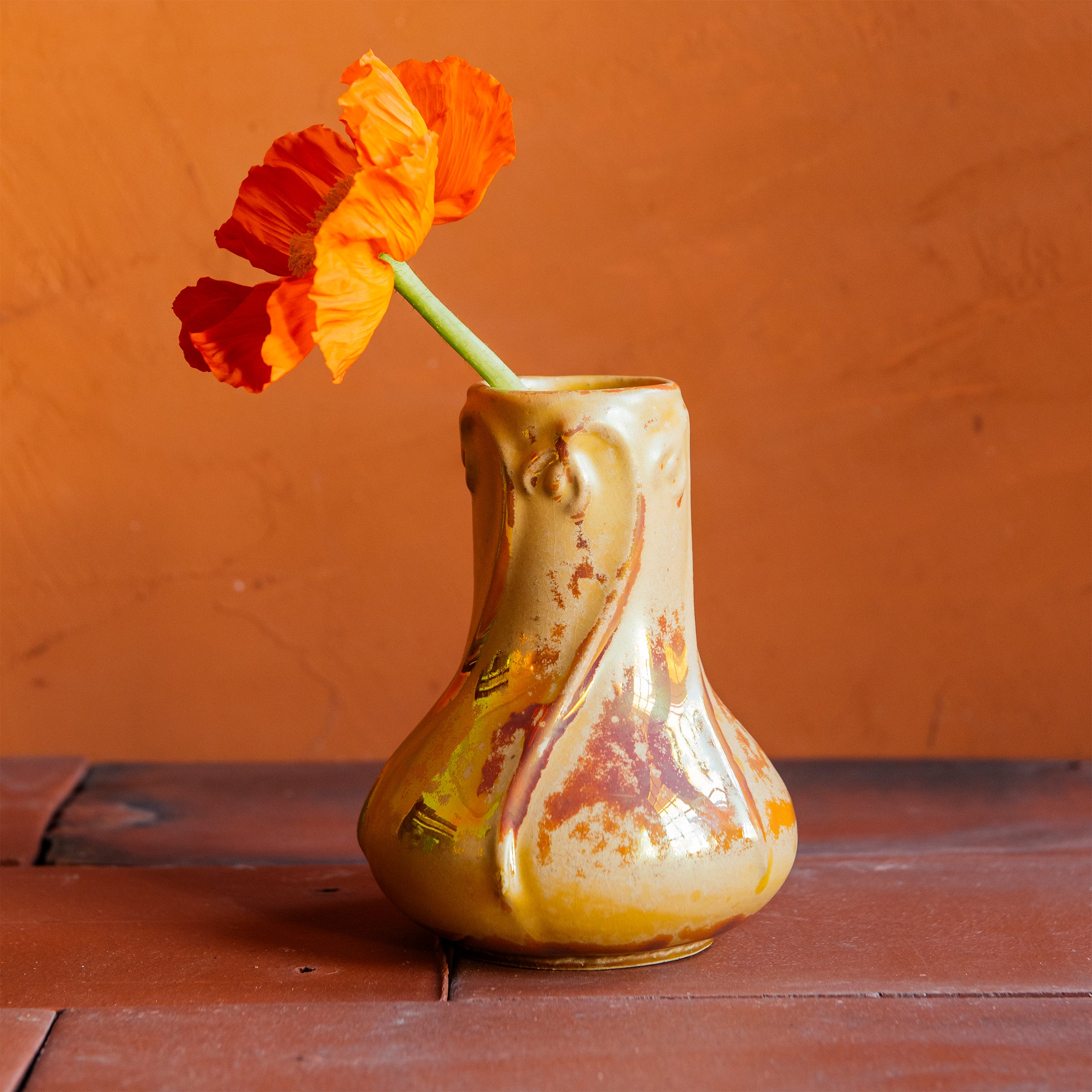 Delicate slip-casted snowdrop floral motifs dance across this Snowdrop Vase in a Sunstone Iridescent glaze that shimmers with the colors of a sunset. The texture of the vase varies with areas that are matte-yellow and reflective features glistening in orange and pinkish hues. The vase rests on terracotta roof tiles with a lime-wash orange background. A single orange poppy is proudly displayed inside of the vase. 