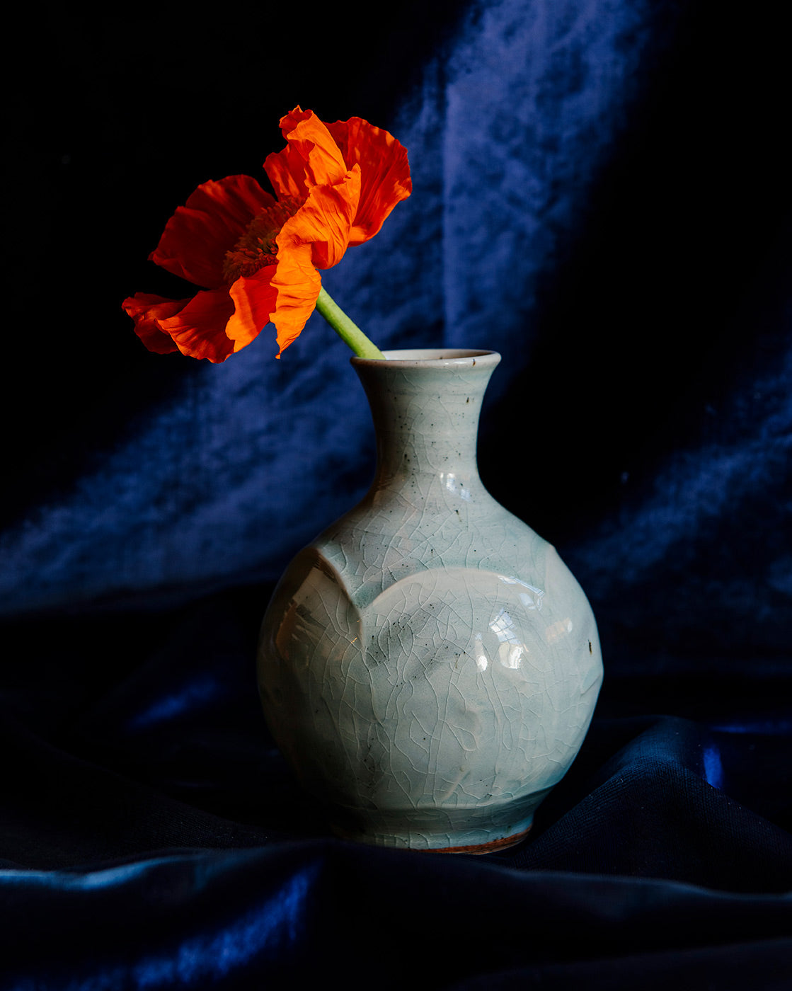 A Pewabic bud vase in a glossy, light-blue Frost glaze set against a blue velvet backdrop and foreground with a single orange poppy flower. 