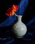 A Pewabic bud vase in a glossy, light-blue Frost glaze set against a blue velvet backdrop and foreground with a single orange poppy flower. 