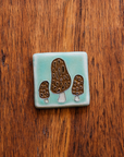 A tile depicting a line-drawn trio of brown Morel mushrooms on a light sea foam-glazed background. 