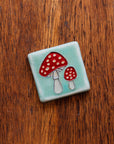 A hand-painted tile depicting a line drawing of a pair of Amanita mushrooms with classic red tops and white spots set on a light sea foam colored background glaze. 