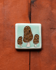 A tile depicting a line-drawn trio of brown Morel mushrooms on a light sea foam-glazed background. 