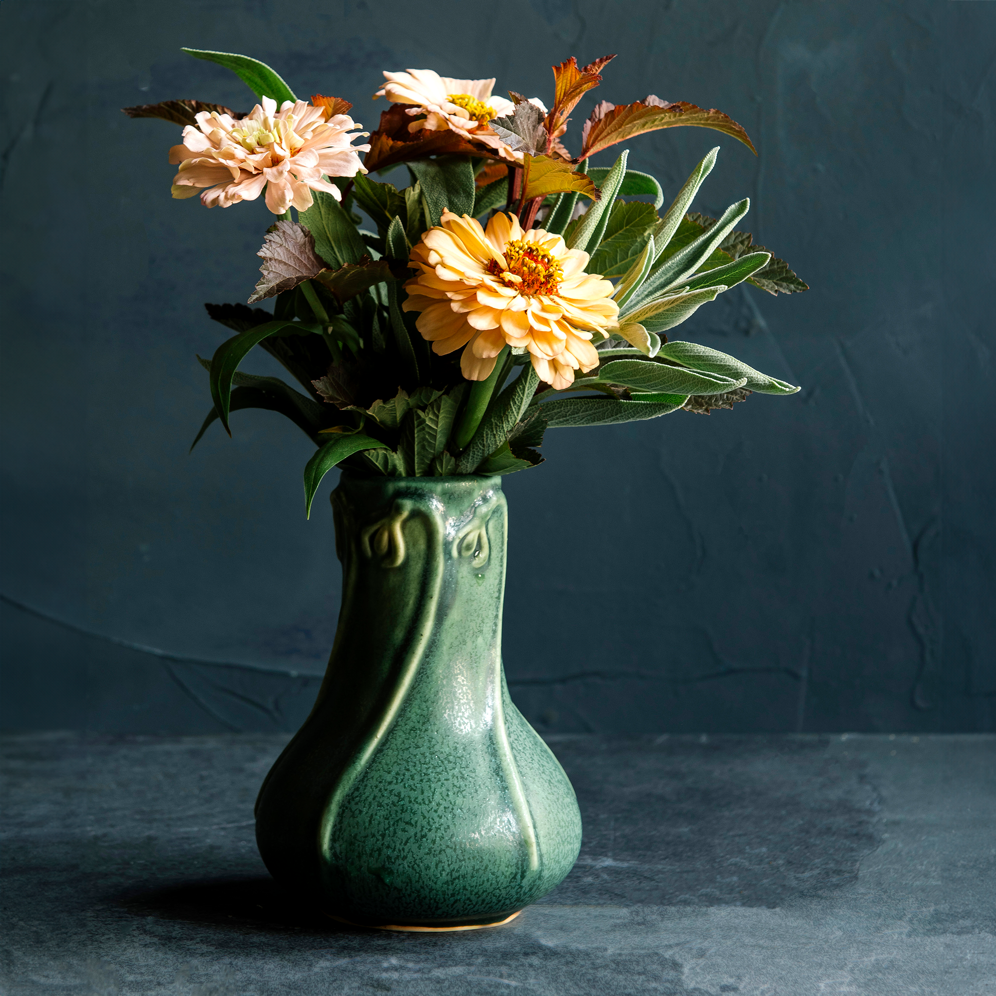 An Elm-glazed ceramic Snowdrop Vase, containing a bouquet of muted peach flowers and sage on a dark background.
