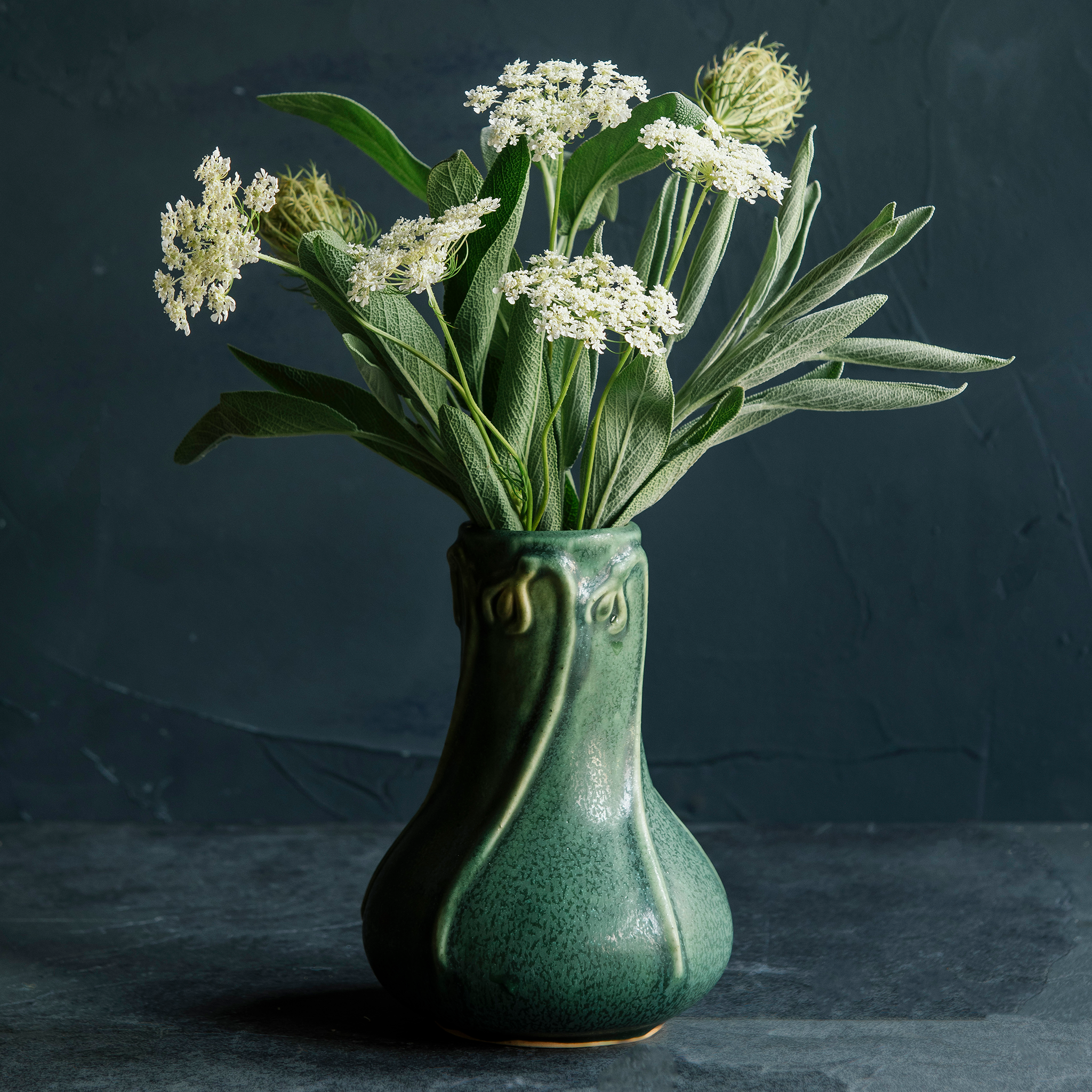 An Elm-glazed ceramic Snowdrop Vase containing a bouquet of white and green flowers, on a dark background.