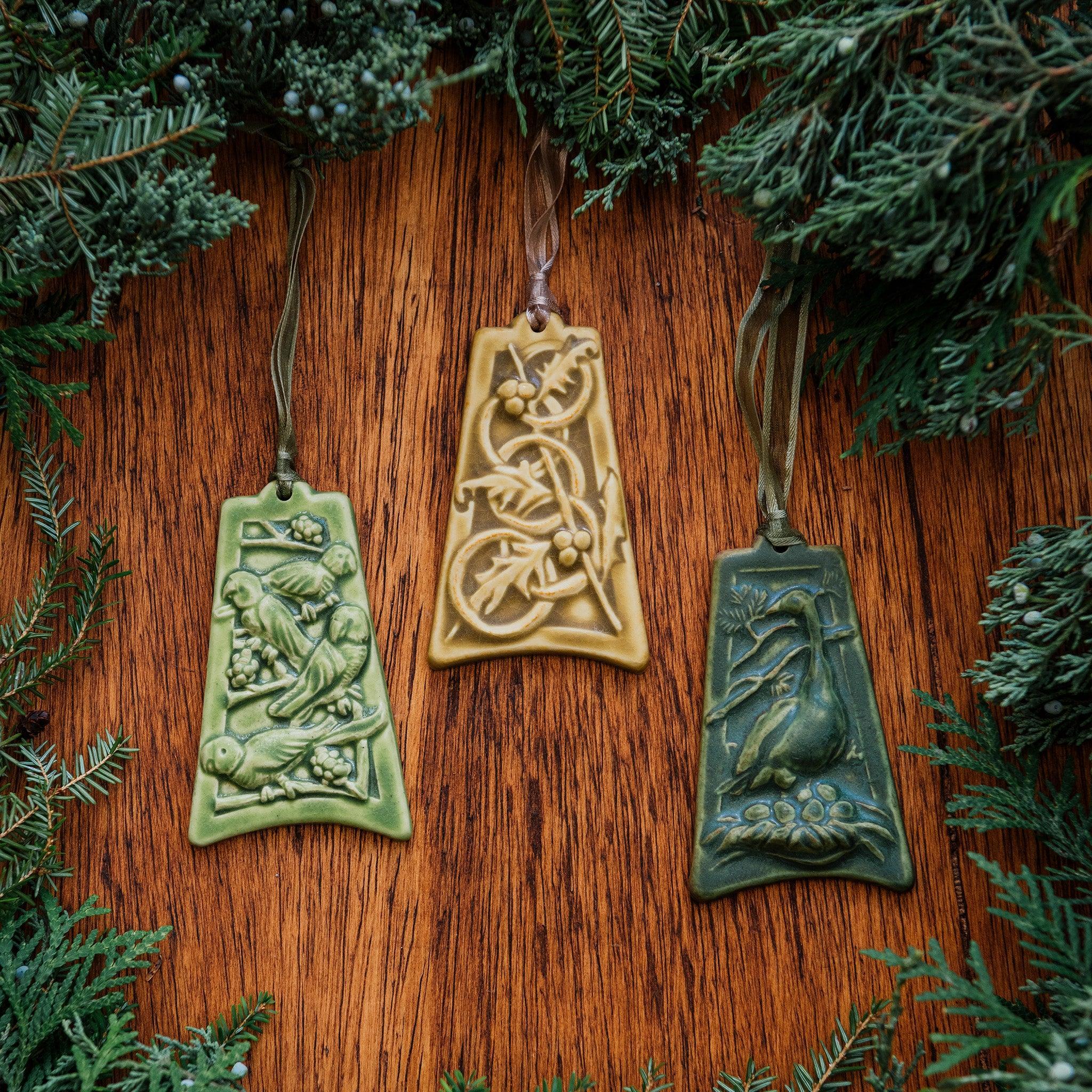 Three colorfully embossed ornaments are lay on a wood background with holiday greenery surrounding them: one has four parrots resting tree branches covered with berries, another has five intertwined rings with holly and berries strewn around them, the last has a goose keeping watch over her nest filled with six eggs.