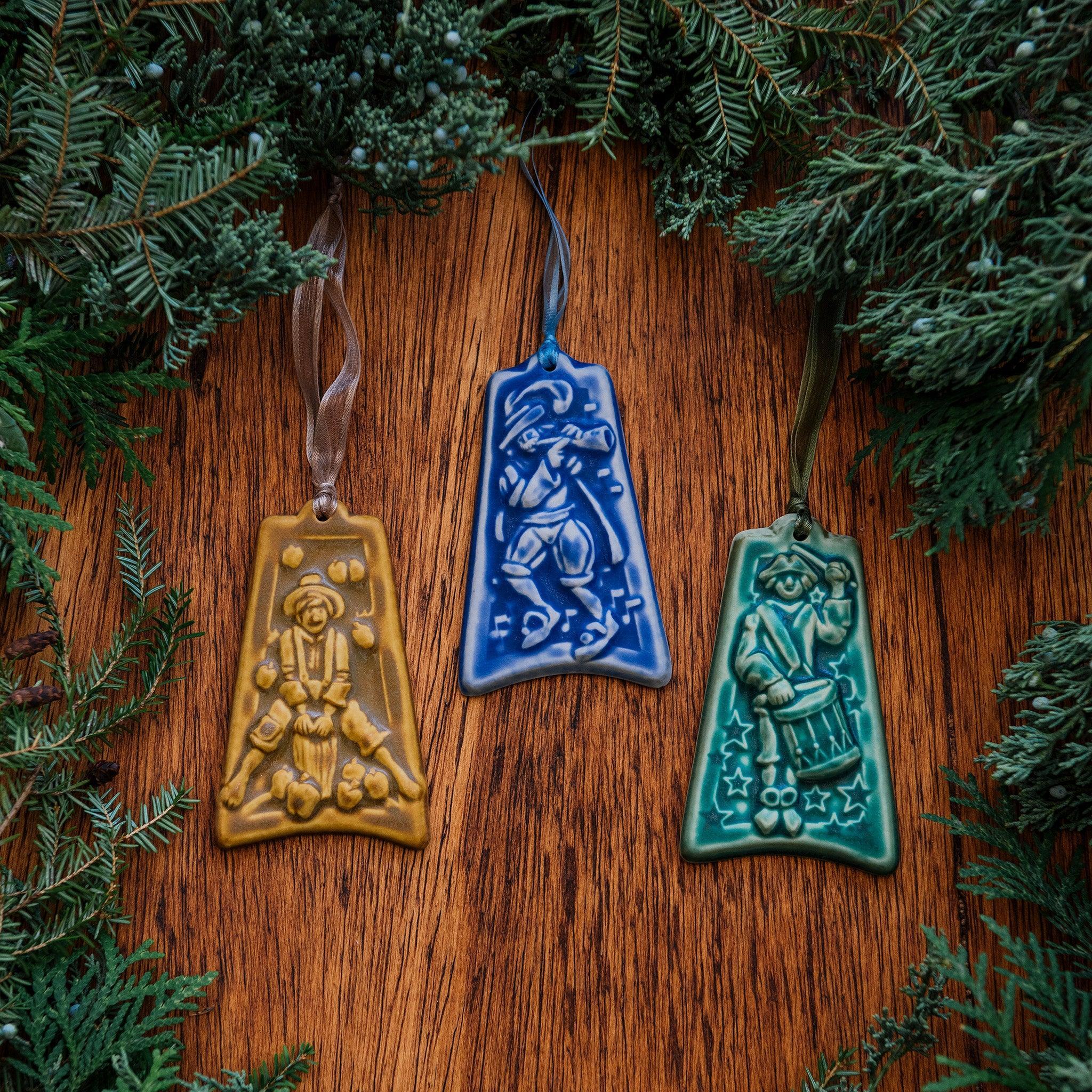 Three colorful embossed ceramic ornaments are on a wood backdrop with holiday greenery surrounding them: one shows a man jumping over a stump surrounded by fallen apples, one shows a man dressed in bloomers and a feathered hat as he plays a horn, the last one has a man dressed in Revolutionary War garb as he marches while playing a drum.  