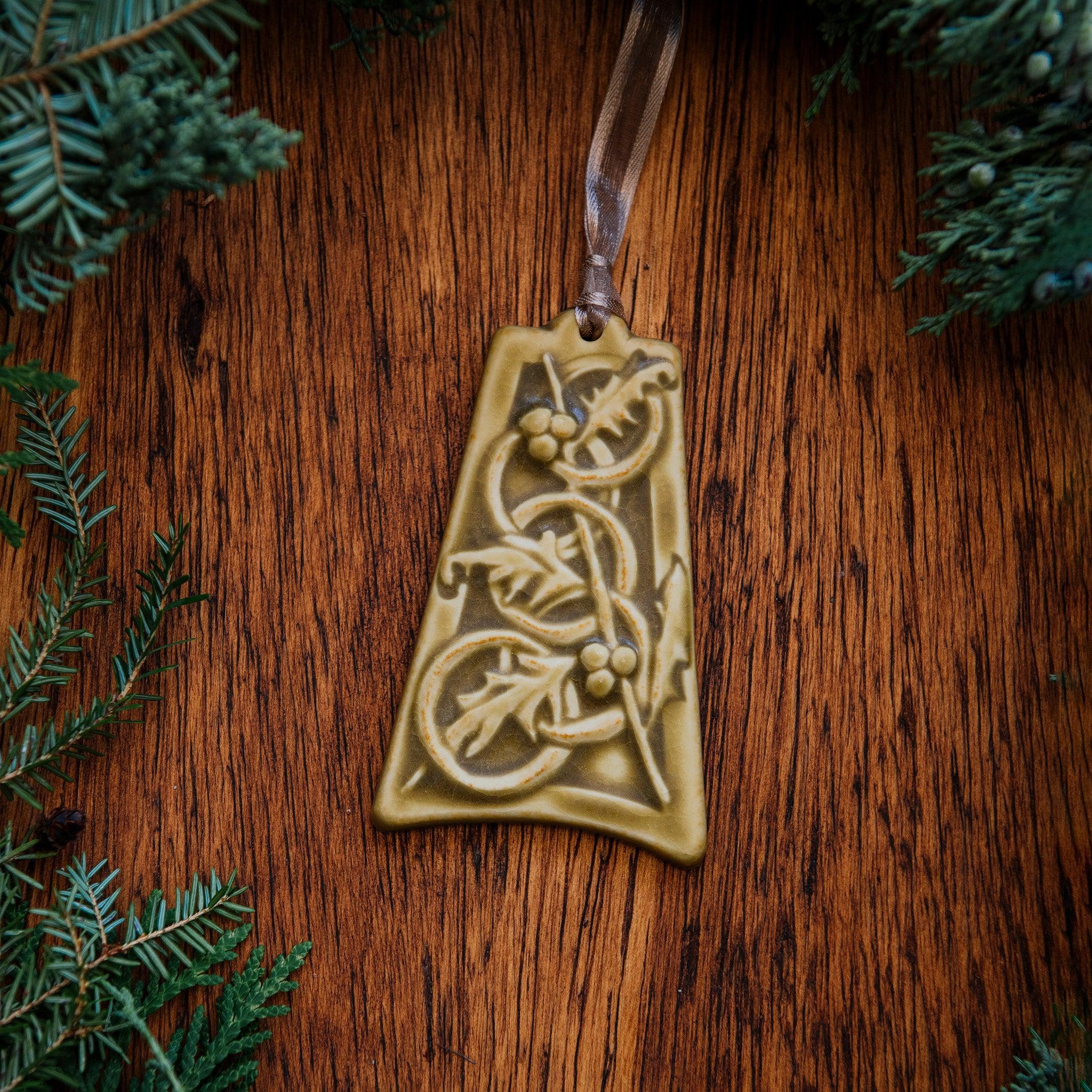 The bell-shaped Five Gold Rings Ornament features five intertwined smooth rings. There are springs of holly leaves and berries throughout the design. The ornament is glazed in a matte yellow glaze.