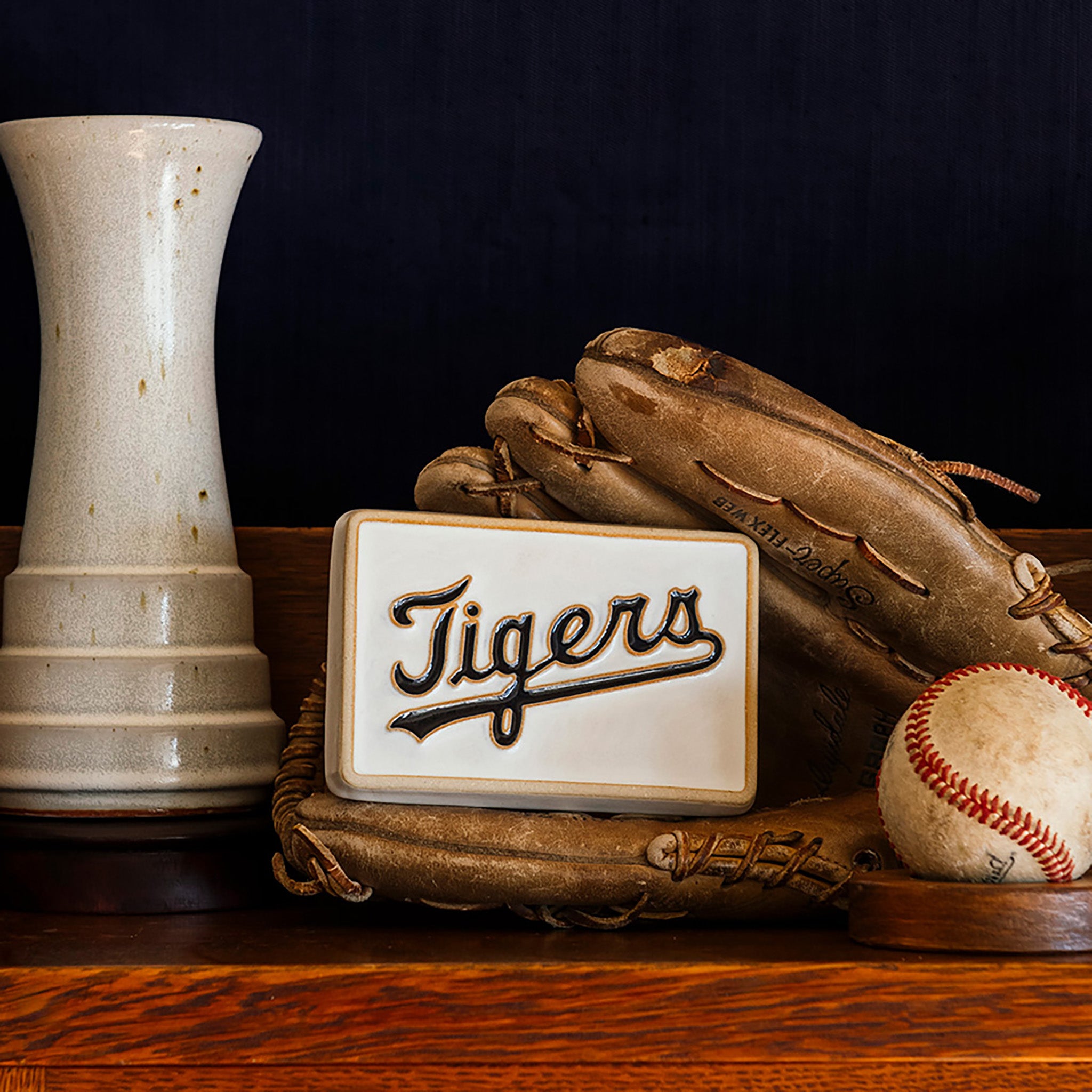 Baseball glove, baseball, Pewabic Step Vase in a Birch glaze, and a 4x6 'Tigers' tile on a wooden shelf.