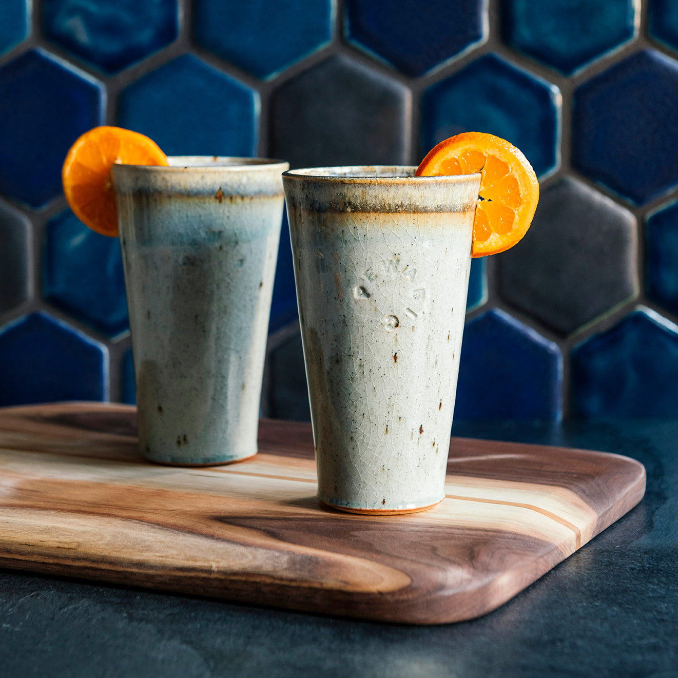Two ceramic Pewabic Pints in a glossy Frost glaze, garnished with orange slices on a wooden cutting board against a blue hex tile background. 