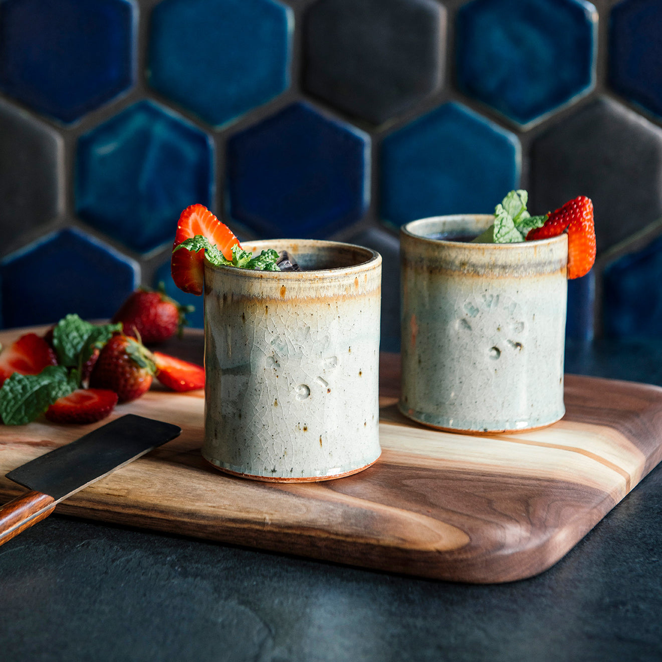 Two ceramic Rocks Cups in a glossy Frost glaze with mint and strawberries on a wooden cutting board against a blue tiled wall.