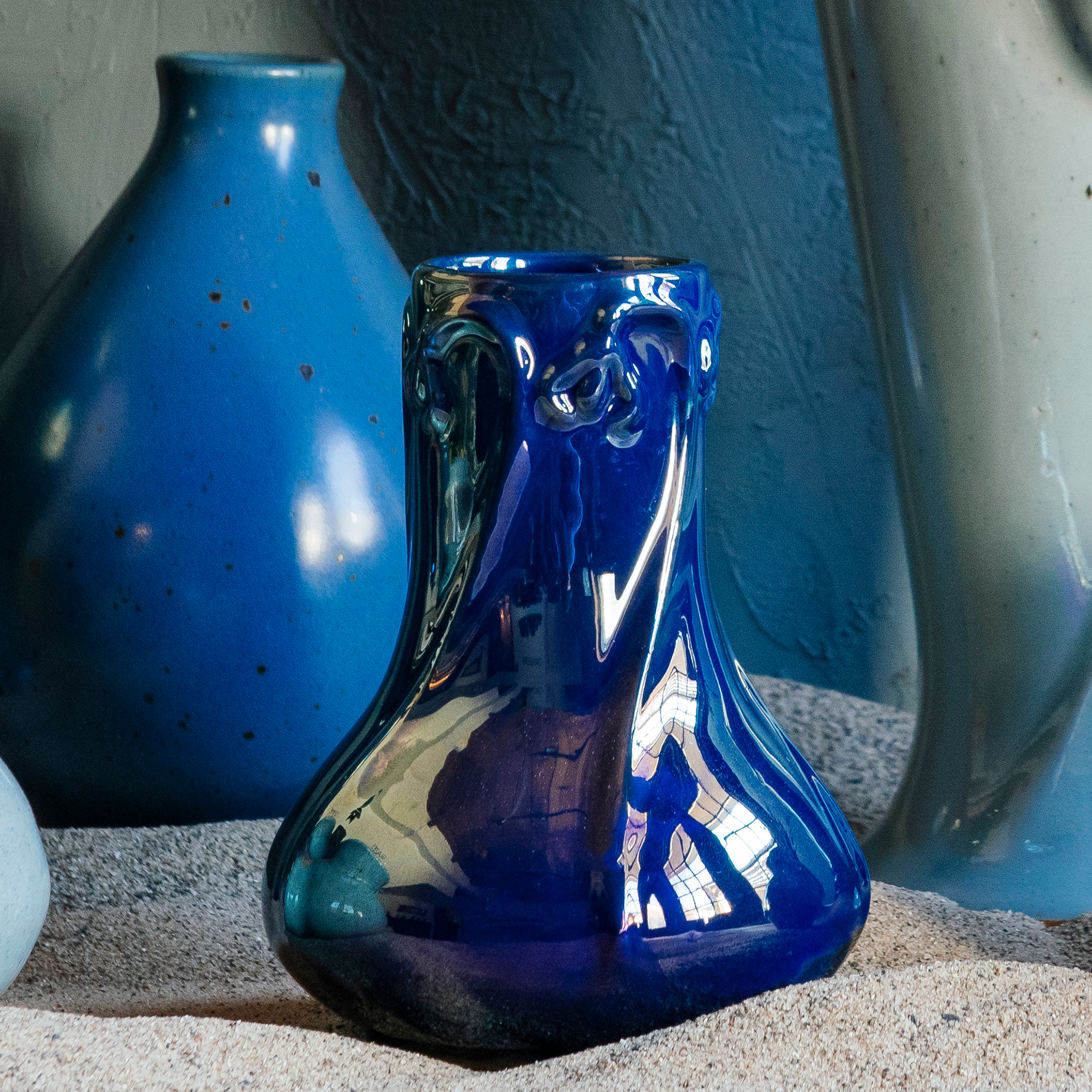 A handcrafted Azurite iridescent Pewabic Snowdrop vase is displayed in a beach setting that includes other Pewabic vases in Frost and Leland Blue in the background.