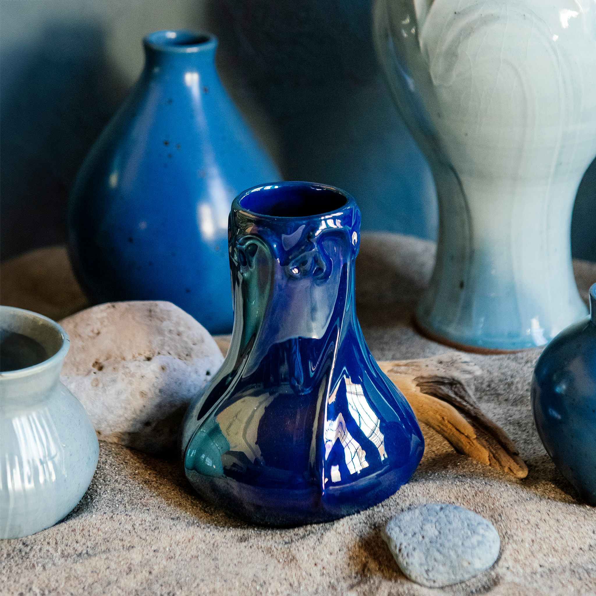 A handcrafted Azurite iridescent Pewabic Snowdrop vase is displayed in a beach setting that includes other Pewabic vases in Frost and Leland Blue in the background.