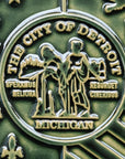 Detail photo of the centerpiece of the Detroit Flag Tile. The circular emblem reads “The City of Detroit” and “Michigan” at the top and bottom. At its center, are two women in red and blue robes. Fire is in the background of the woman on the left and reads “Speramus Meliora” which is latin for “We hope for better things”. The woman on the left is surrounded by a cityscape and reads “Resurget Cineribus” which is “It will rise from the ashes” in latin. 