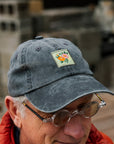Man wearing a dark gray cap with a floral patch and sunglasses, outdoors.