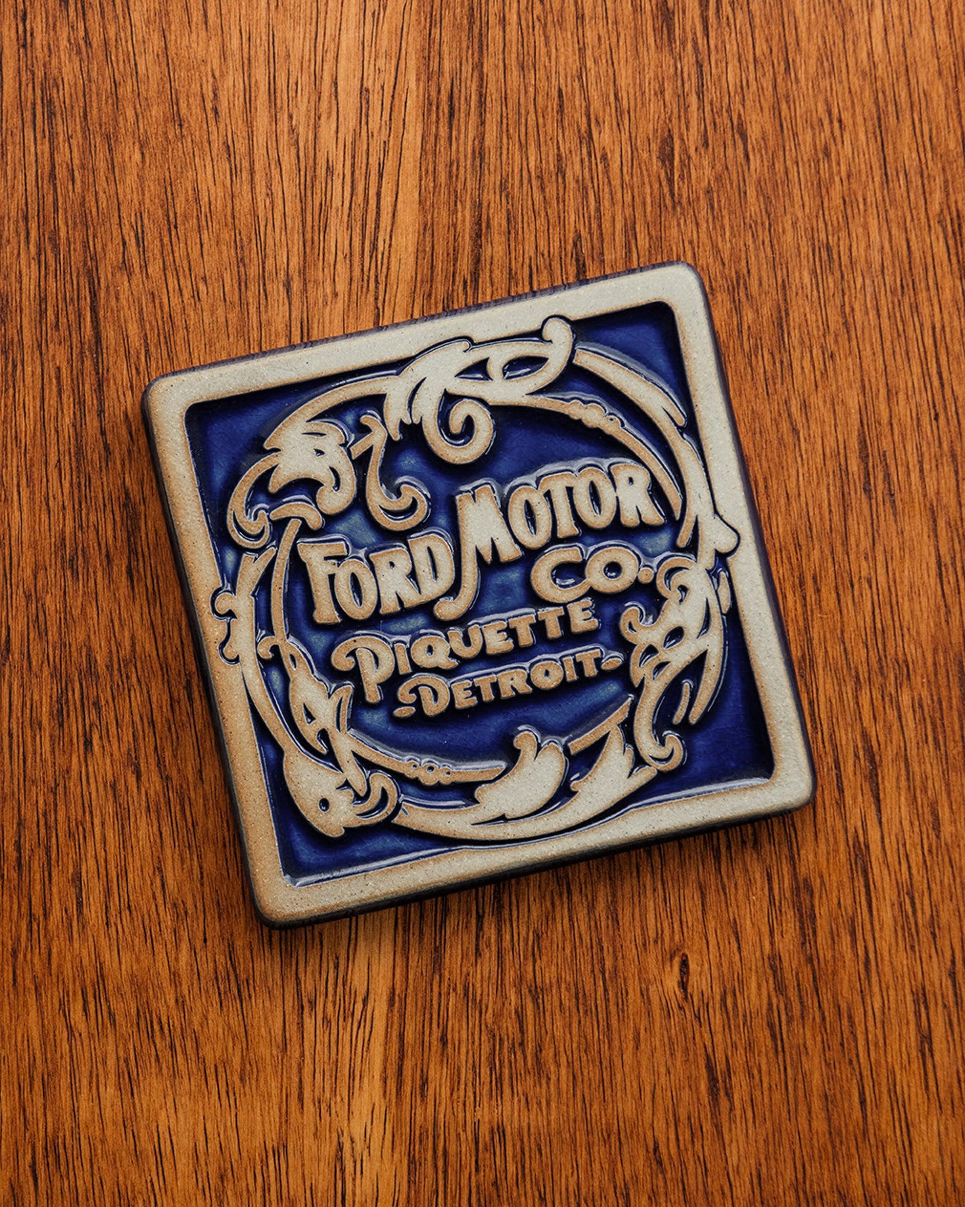 Ford Motor Co. Piquette logo tile on a warm wooden background.