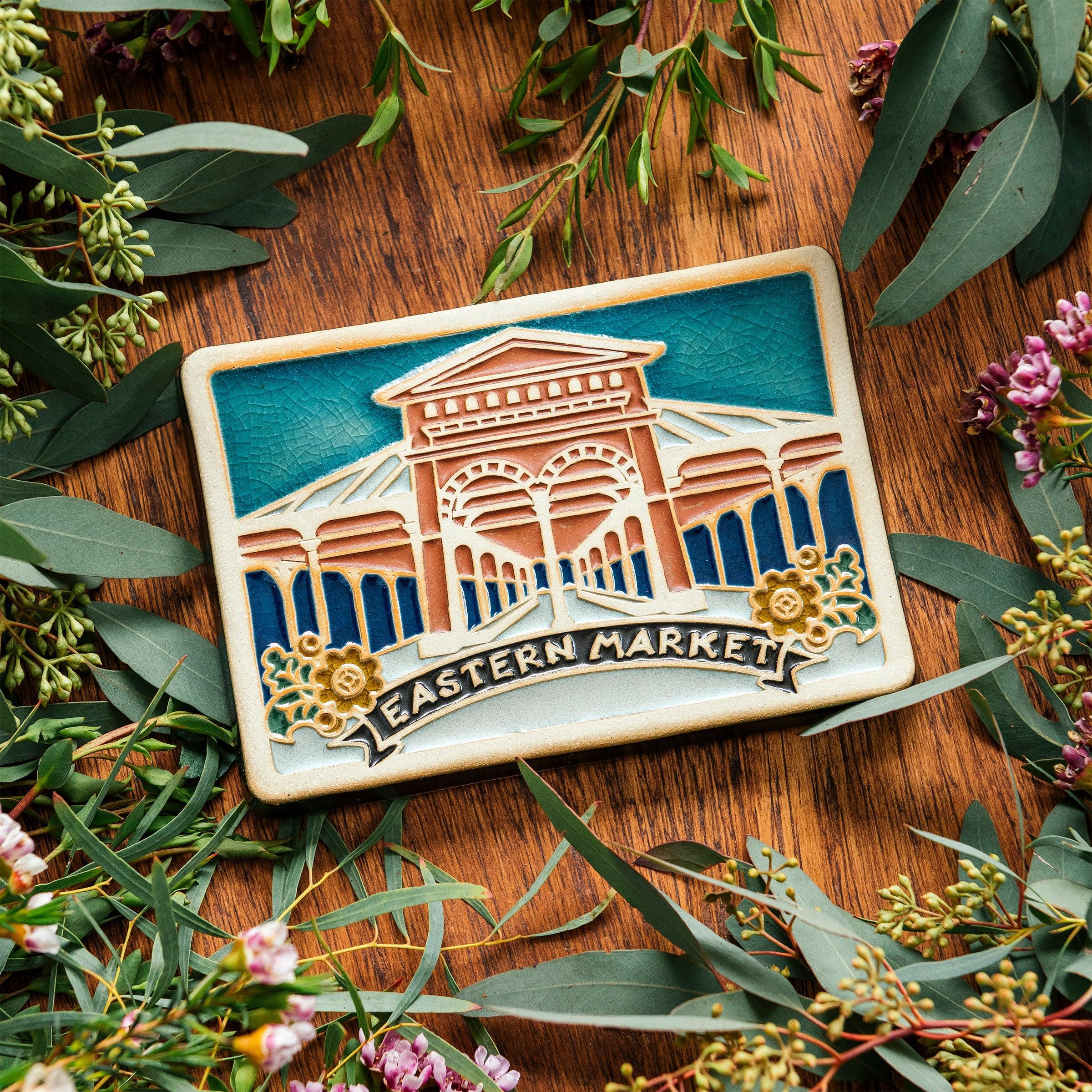 A hand-painted Eastern Market Postcard Tile rests on a wooden table with a warm finish surrounded by dainty wax flowers and seeded eucalyptus. 