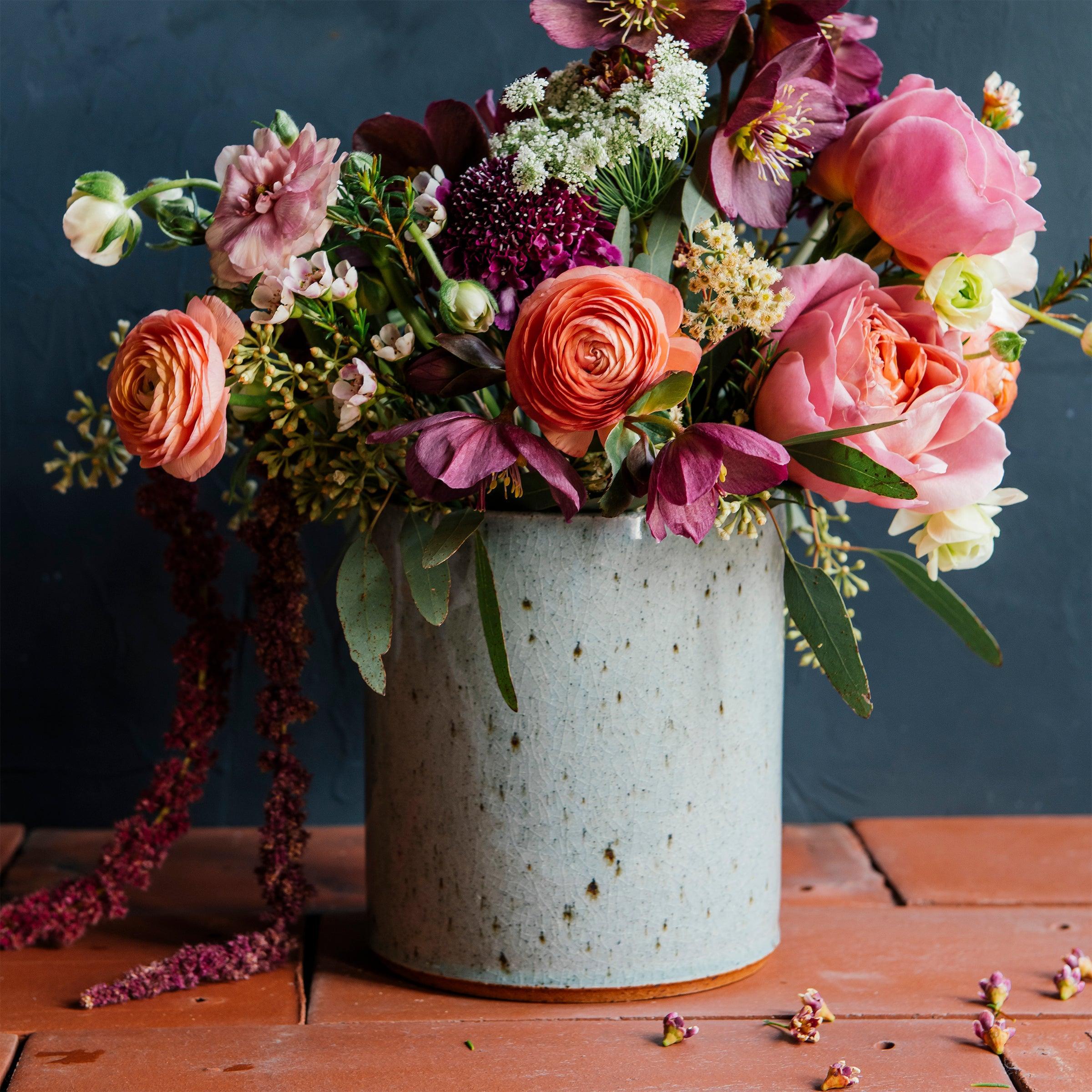 A Pewabic Crock featured in the icy-blue Frost glaze. Dark iron flecks are visible on the glossy surface of the vessel. The crock is set on terracotta roof tiles with a lime-wash backdrop and brilliant summer florals, including garden roses. 