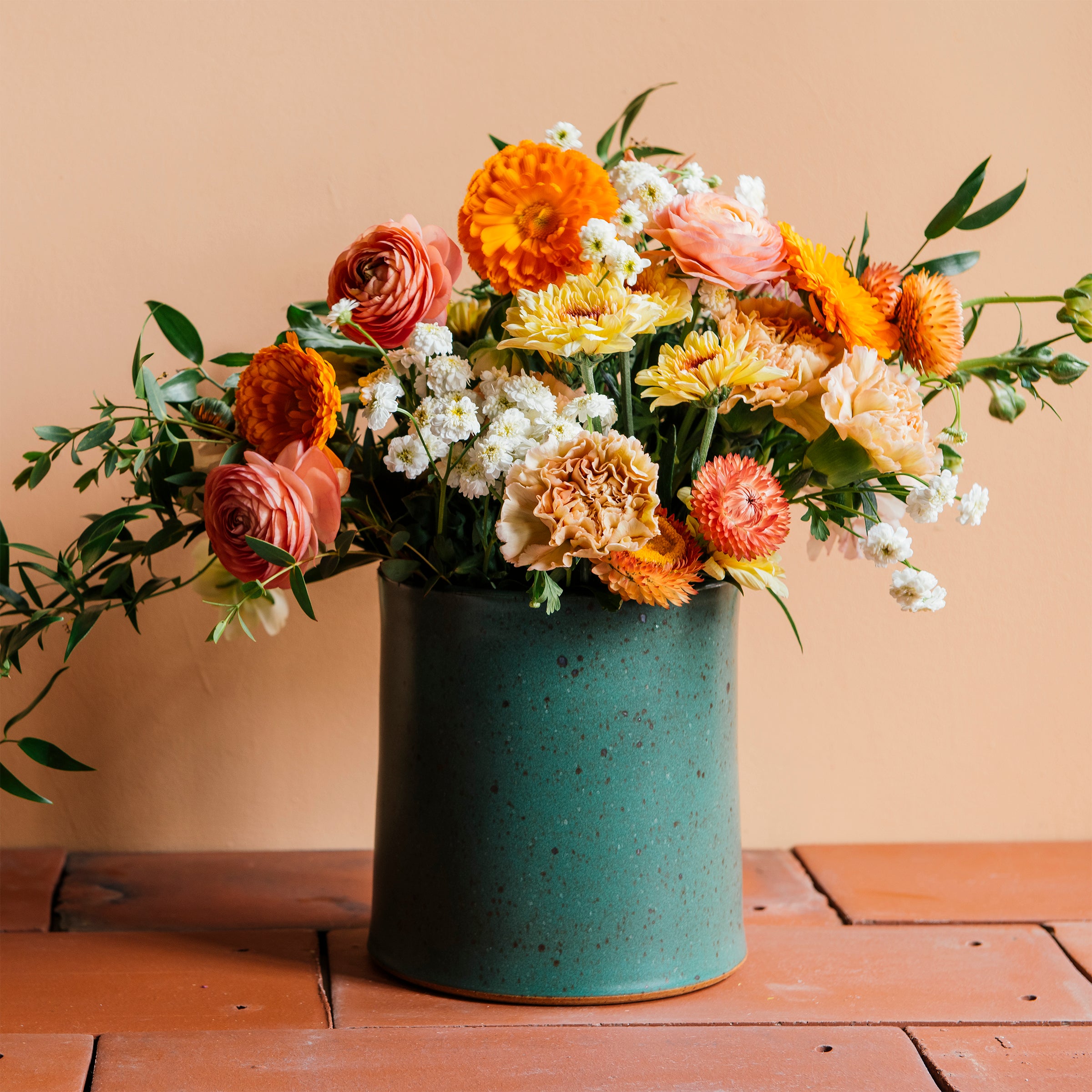 A colorful orange and blush flower arrangement in a Pewabic Crock featured in the matte Greenstone glaze, complete with visible iron flecks on its surface. The vessel is set upon terracotta roof tiles and a light peach background. 